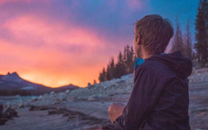 a young person looks away from the camera in the direction of a sunset behind mountains in yosemite